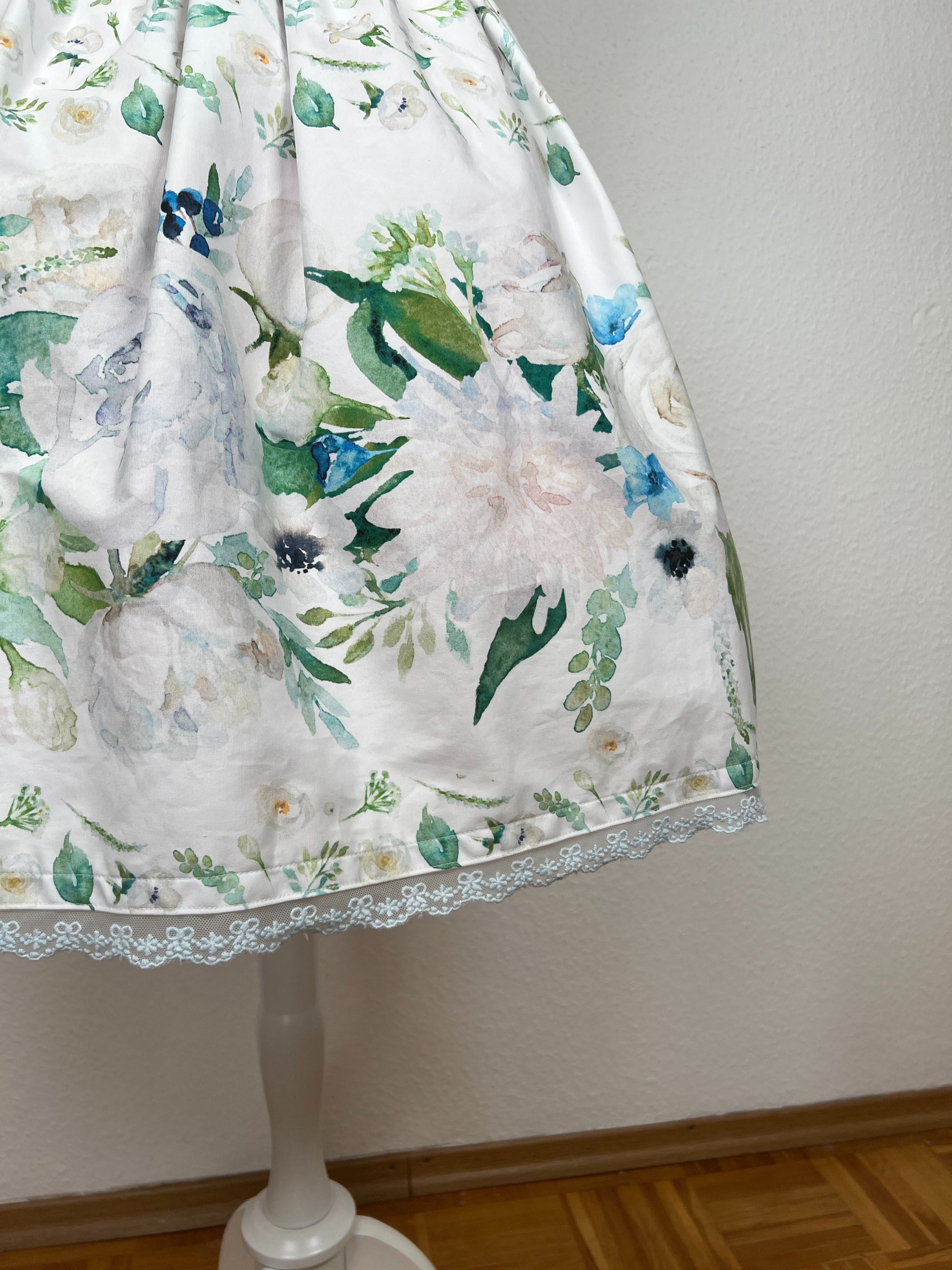 Detailansicht Rock, Bordüre mit großen cremefarbenen und hellblauen Blumen und grünen Blättern auf weißem Grund, hellblaue Spitze am Saum