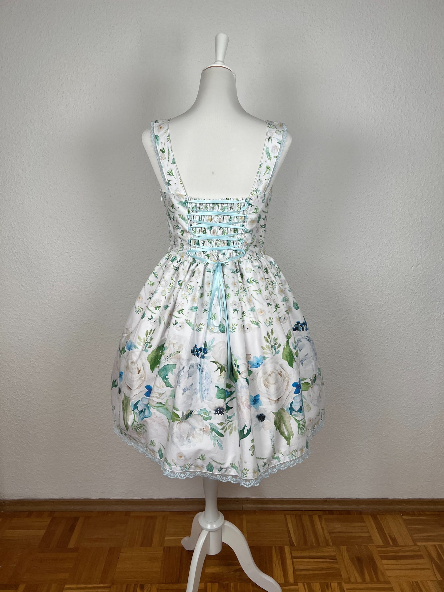 Rückansicht Kleid mit Trägern und gerüschtem Rock, weißer Stoff mit cremefarbenen Blumen, Bordüre mit großen Blumen in creme und blau, blaue Schnürung am Rücken, blaue Spitze an Saum und Trägern