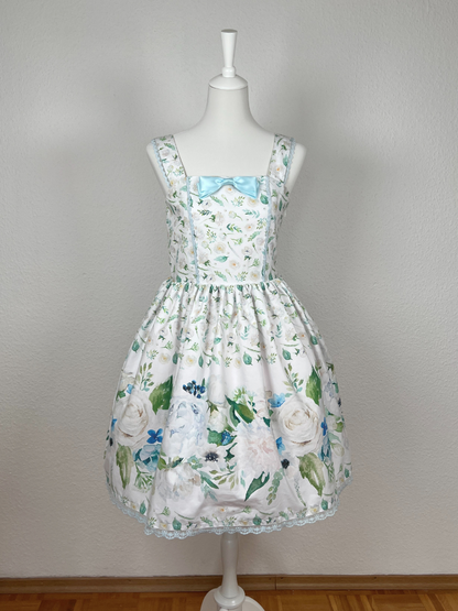 Kleid mit Trägern und gerüschtem Rock, weißer Stoff mit cremefarbenen Blumen, Bordüre mit großen Blumen in creme und hellblau, blaue Schleifenbrosche, blaue Spitze an Saum, Oberteil und Trägern