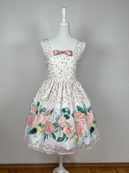 Kleid mit Trägern und gerüschtem Rock, weißer Stoff mit rosa Blumen, Bordüre mit großen Blumen in creme und rosa, rosa Schleifenbrosche, rosa Spitze an Saum, Oberteil und Trägern