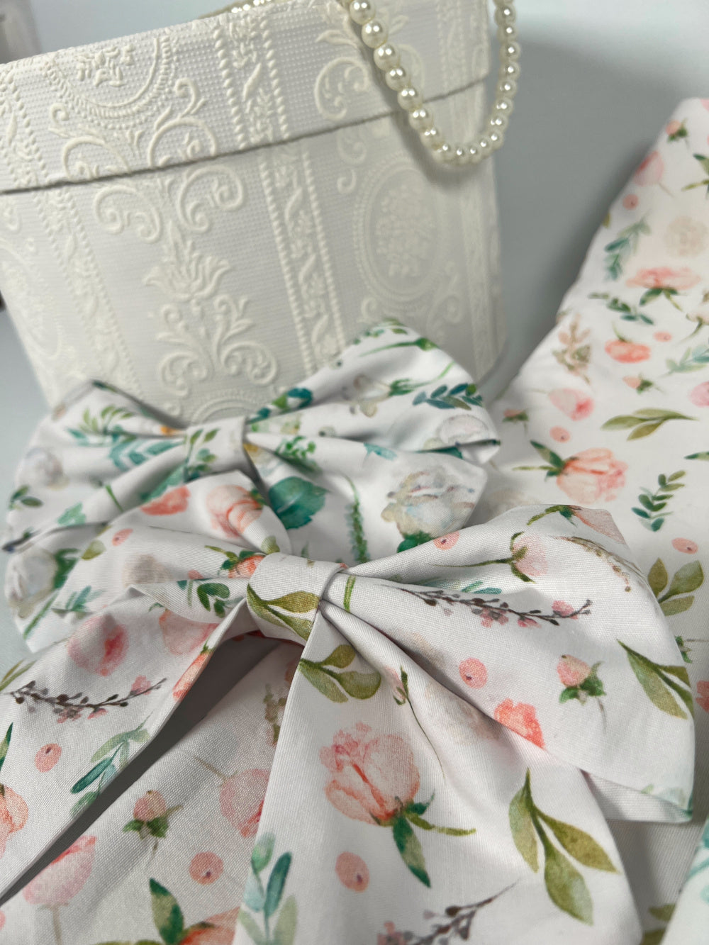 zwei Schleifen mit Blumenmuster im Vordergrund, weiße Hutschachtel mit barockem Muster im Hintergrund, auf ihr eine Perlenkette